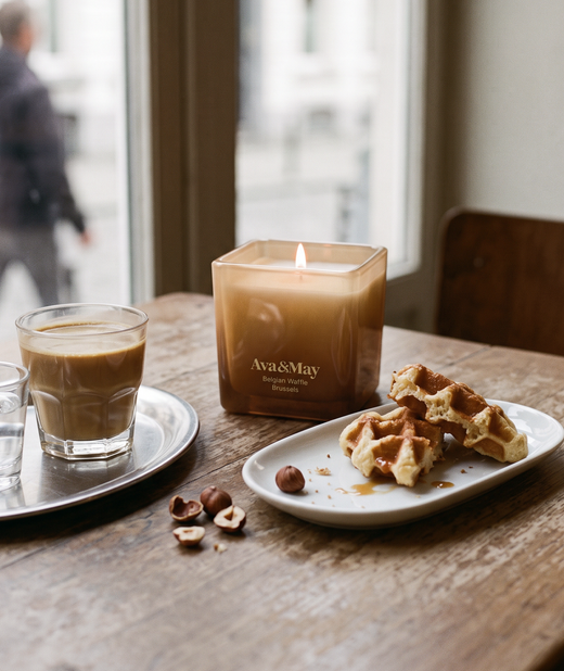 Die brennende Ava&May Belgian Waffle Kerze (220 Gramm) in einem quadratischen Glasgefäß auf einem Holztisch neben einem Glas Kaffee, einigen Haselnüssen und einer süßen belgischen Waffel