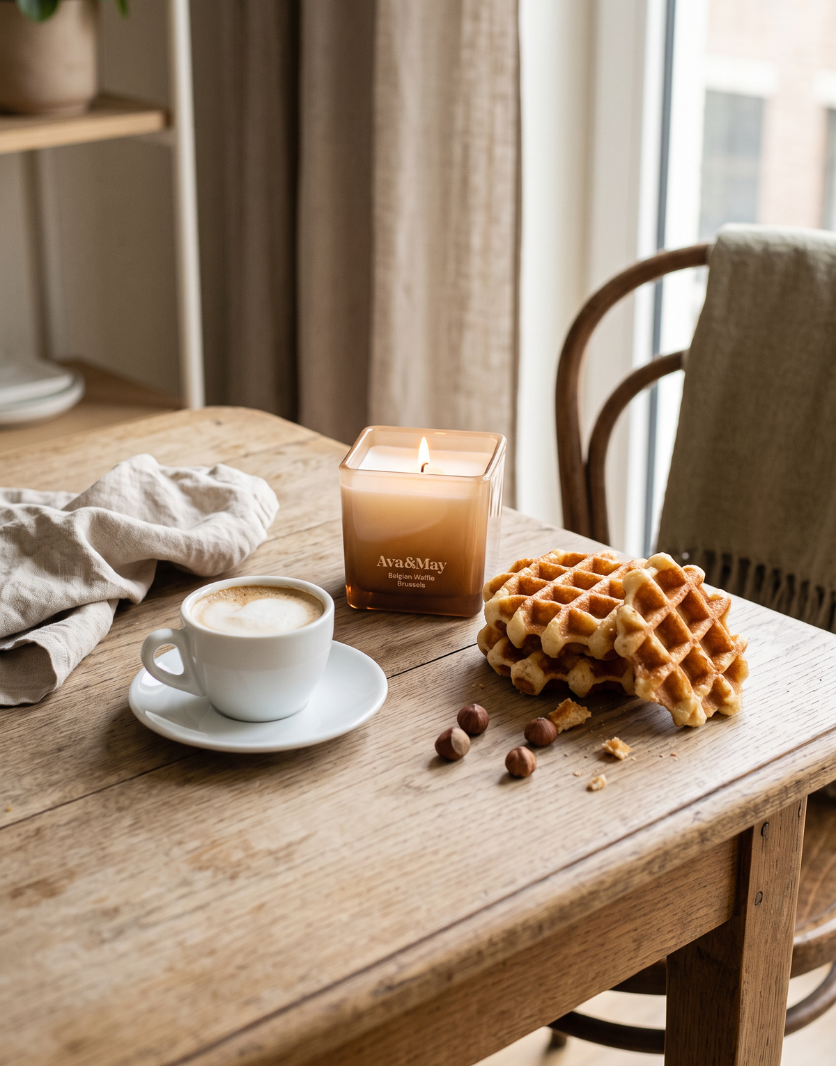 Die brennende Kerze Ava&May Belgian Waffle (220 Gramm) steht auf einem Holztisch in einem Lokal, umgeben von einer Tasse Cappuccino mit cremigem Schaum, Haselnüssen und belgischen Waffeln.