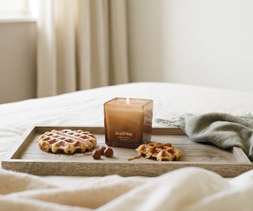 Die brennende Kerze „Ava&May Belgian Waffle“ (220 Gramm) steht auf einem Holztablett, das auf einer Tischdecke liegt, umgeben von Waffelgebäck und Haselnüssen.