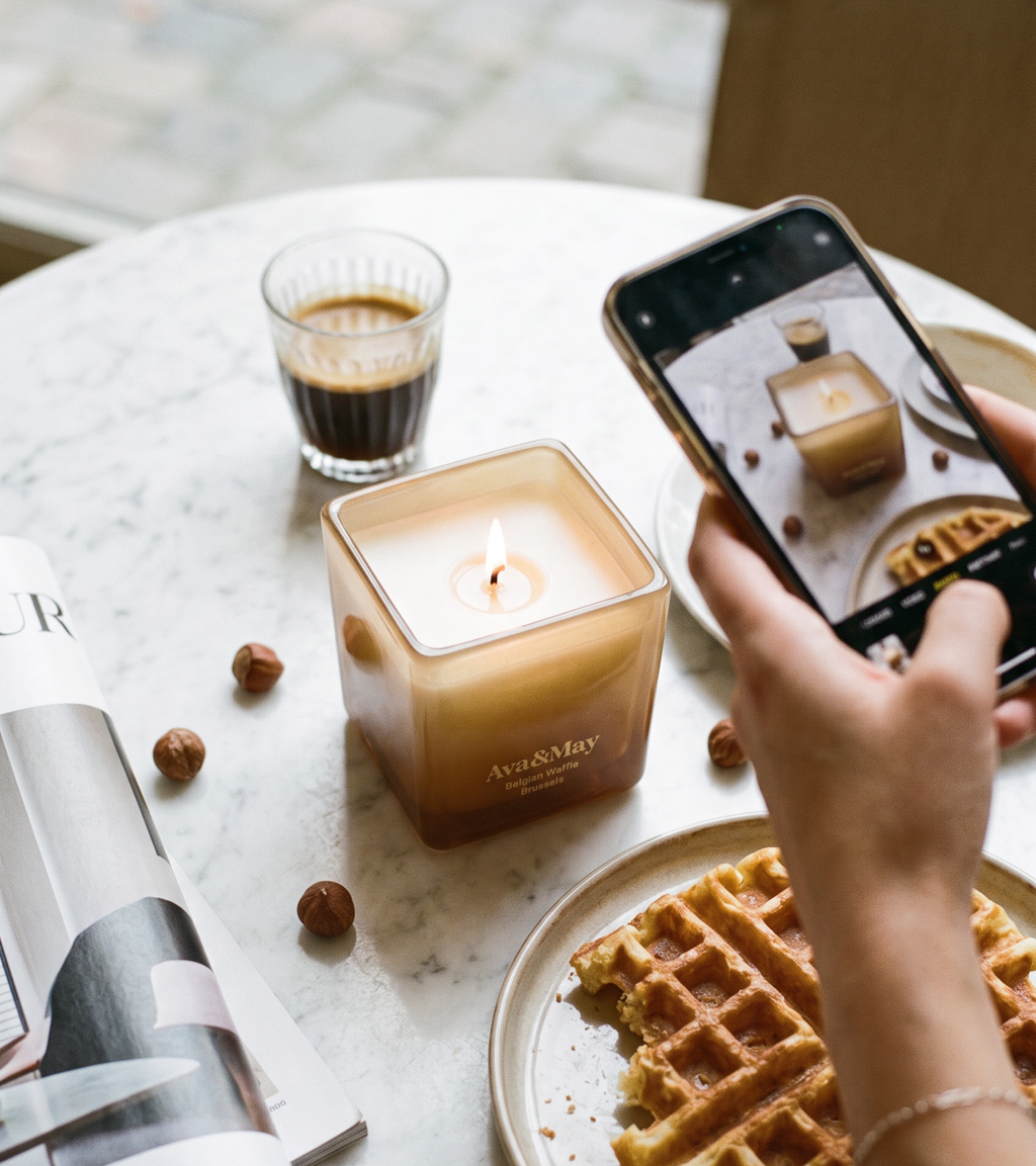 Die Ava&May Kerze Belgian Waffle (220 Gramm) brennt auf einem Marmortisch, umgeben von einem Espresso, einer belgischen Waffel, einer Einrichtungszeitschrift und Haselnüssen, während sie von einer Person mit einem Handy in der Hand fotografiert wird