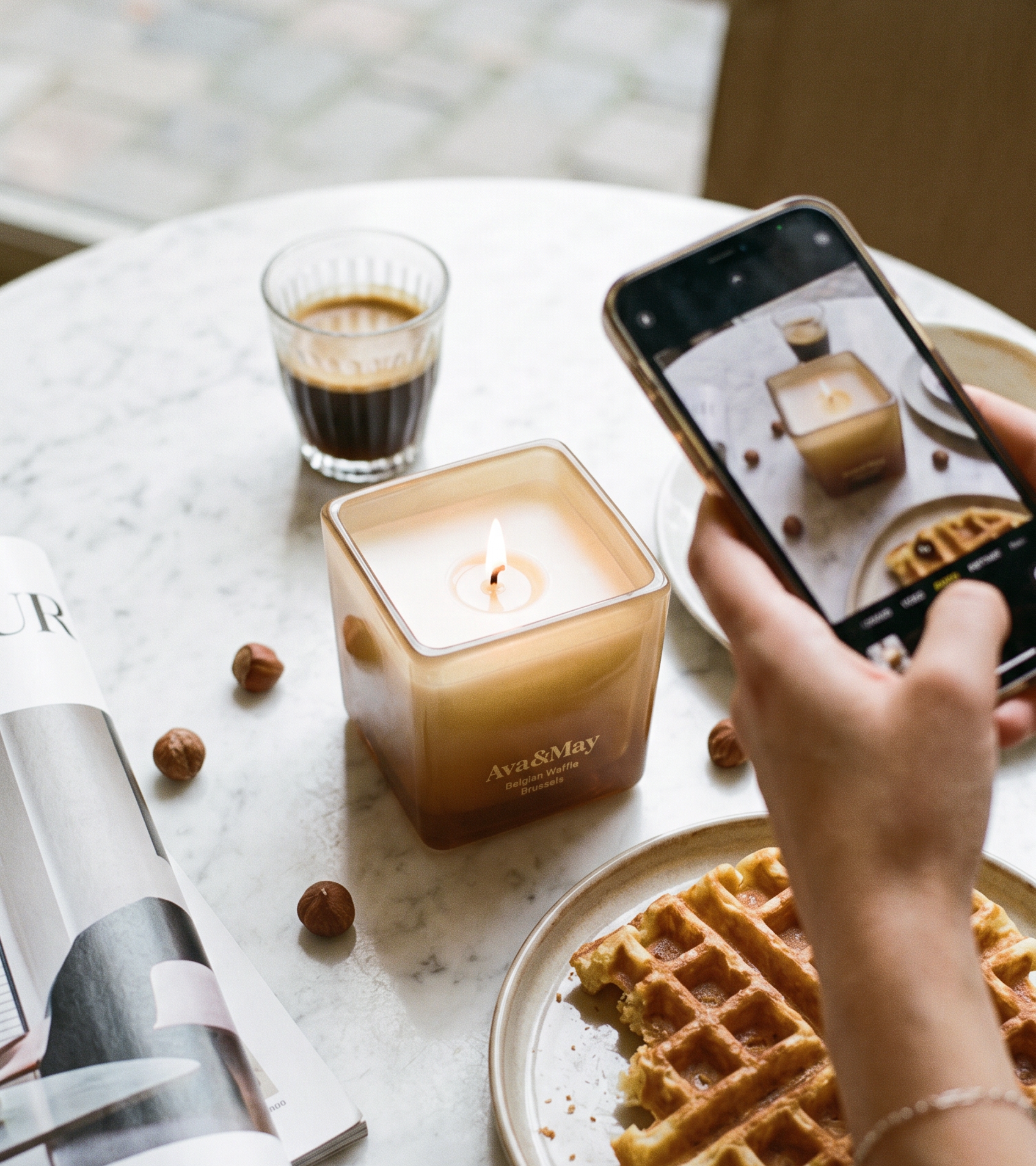 Die Ava&May Kerze Belgian Waffle (220 Gramm) brennt auf einem Marmortisch, umgeben von einem Espresso, einer belgischen Waffel, einer Einrichtungszeitschrift und Haselnüssen, während sie von einer Person mit einem Handy in der Hand fotografiert wird