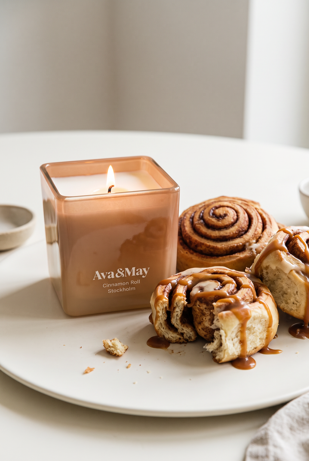 Die Kerze Cinnamon Roll (220 Gramm) von Ava&May brennt auf einem Keramiktellerchen, daneben liegen würzige Zimtschnecken, die mit Karamellcreme überzogen sind