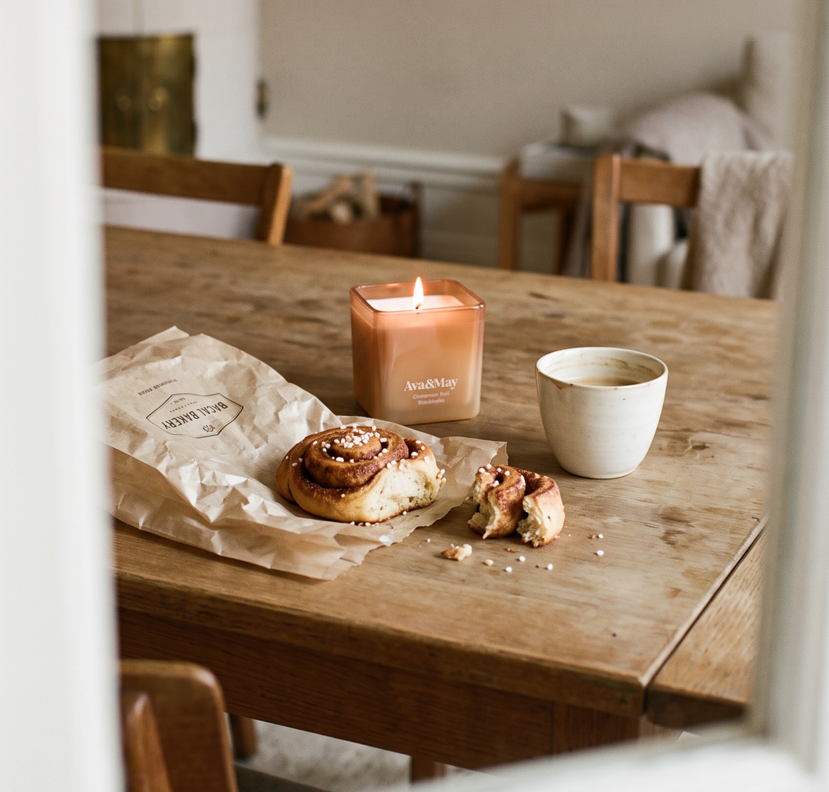 Die 220-Gramm-Kerze Cinnamon Roll von Ava&May brennt auf einem Holztisch in einem Esszimmer, daneben stehen eine Tasse Kaffee und ein Zimtgebäck aus einer Bäckerei