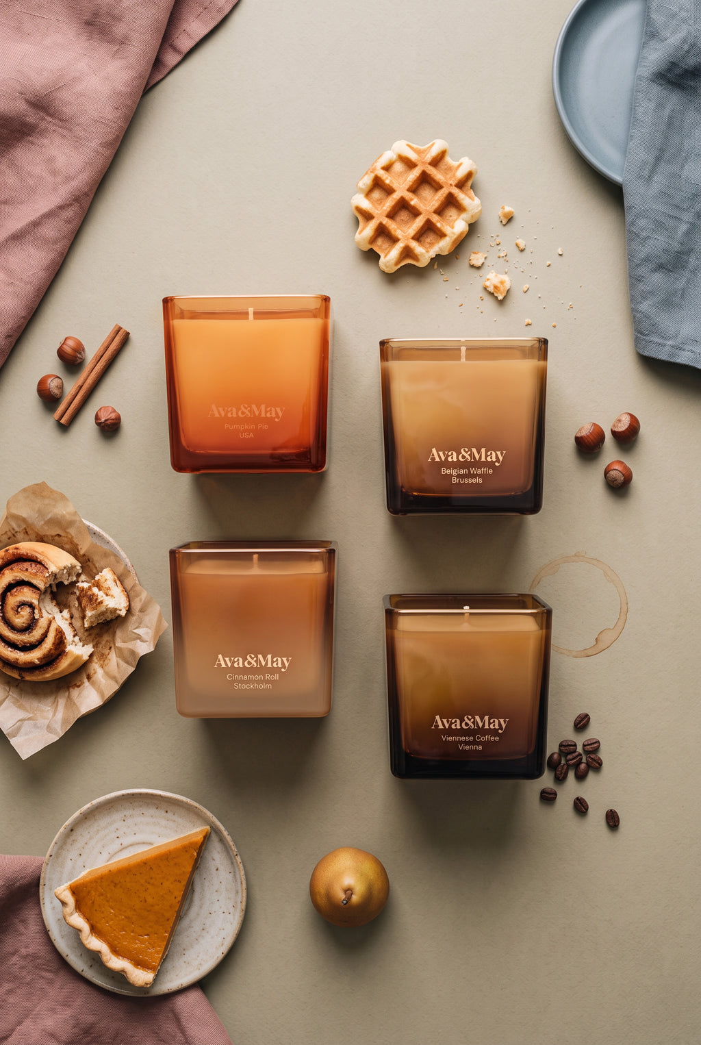 Ava&May Sweet Sensations Kerzenset: Kerzen in den Duftrichtungen Belgian Waffle 220 Gramm, Viennese Coffee 220 Gramm, Pumpkin Pie 220 Gramm und Cinnamon Roll 220 Gramm liegen auf einem grünen Tisch, und daneben stehen ein Kürbiskuchen, ein Zimtgebäck, Zimt, eine belgische Waffel, Haselnüsse und Kaffeebohnen