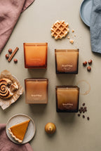 Ava&May Sweet Sensations Kerzenset: Kerzen in den Duftrichtungen Belgian Waffle 220 Gramm, Viennese Coffee 220 Gramm, Pumpkin Pie 220 Gramm und Cinnamon Roll 220 Gramm liegen auf einem grünen Tisch, und daneben stehen ein Kürbiskuchen, ein Zimtgebäck, Zimt, eine belgische Waffel, Haselnüsse und Kaffeebohnen