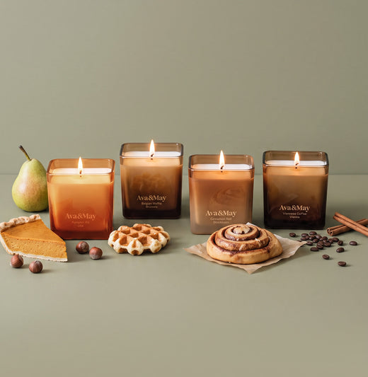 Ava&May Sweet Sensations Kerzenset: Kerzen in den Duftrichtungen Belgian Waffle 220 Gramm, Viennese Coffee 220 Gramm, Pumpkin Pie 220 Gramm und Cinnamon Roll 220 Gramm stehen in einer Reihe, und daneben liegen kleine Leckereien und Gebäckstückchen, eine Birne, Zimtstangen, Kaffeebohnen und Haselnüsse.
