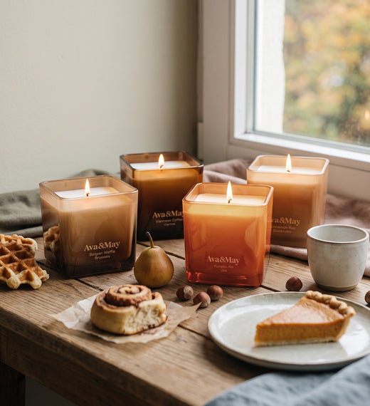 Ava&May Sweet Sensations Kerzenset: Kerzen in den Duftrichtungen Belgian Waffle 220 Gramm, Viennese Coffee 220 Gramm, Pumpkin Pie 220 Gramm und Cinnamon Roll 220 Gramm, die auf einem Holztisch zusammen mit Waffeln, Kürbiskuchen und Zimtschnecken brennen