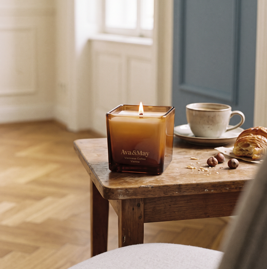 Eine brennende Kerze Ava&May Viennese Coffee auf einem Holztisch mit einer Tasse Kaffee, einem Croissant und Haselnüssen
