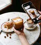 Ava&May-Kerze Viennese Coffee 220 Gramm, brennend, mit einer Tasse Kaffee, einem Croissant und einer Hand, die sie mit einem Handy fotografiert